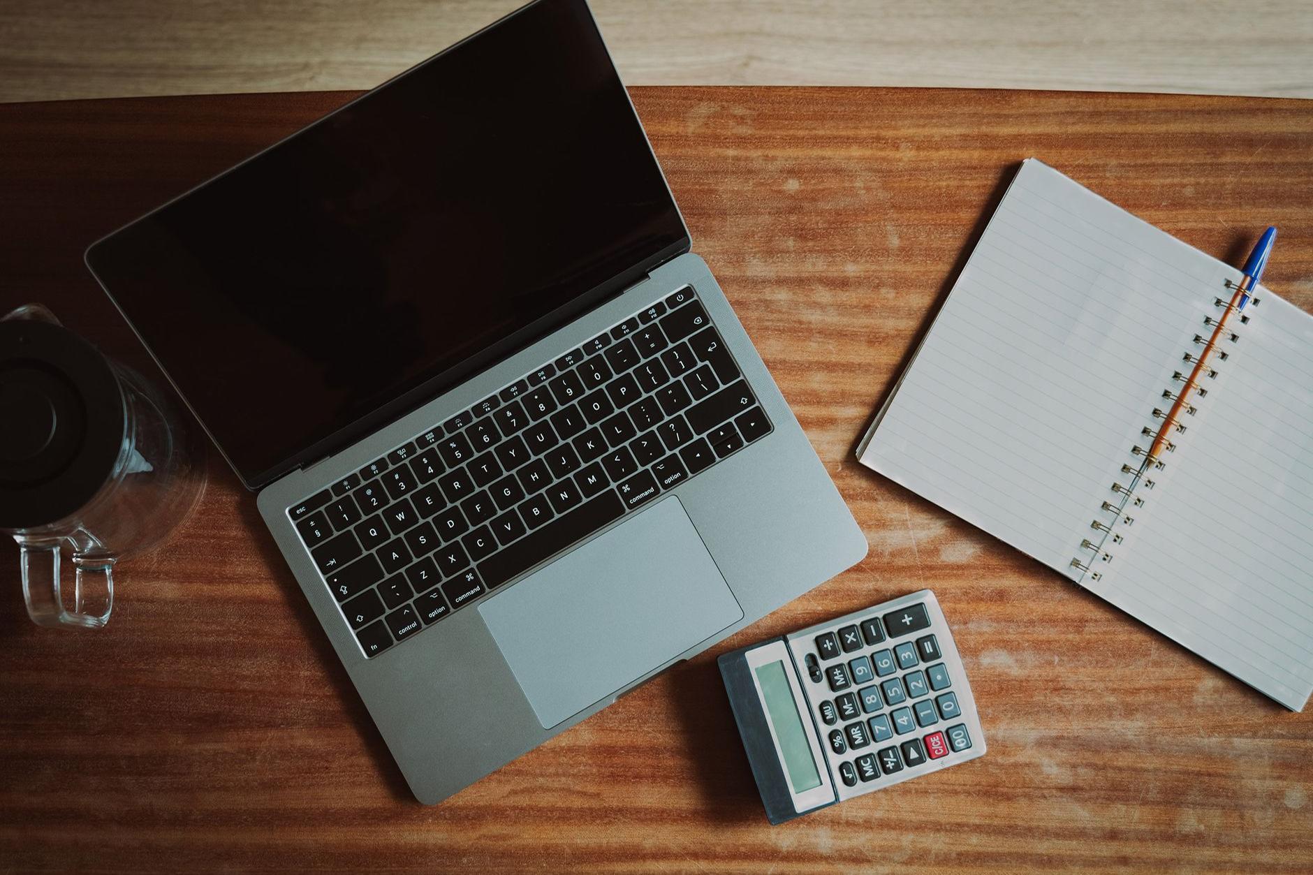A laptop, open lined notebook, pen and calculator arranged on a wooden desk.